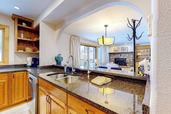 A kitchen with granite countertops, wooden cabinets, a double sink, and a dining area with a stone fireplace. - Mountain-view condo with fireplace, W/D, & shared amenity access (Vail)