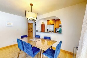 A dining area with a wooden table and blue chairs, a kitchen with wooden cabinets, and a ceiling light fixture. - Mountain-view condo with fireplace, W/D, & shared amenity access (Vail)