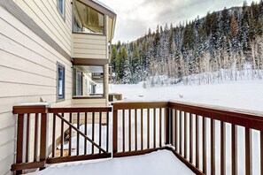 A wooden deck with a snow-covered ground, a two-story building with large windows, and a forested background. - Mountain-view condo with fireplace, W/D, & shared amenity access (Vail)
