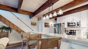 Una cocina moderna con armarios blancos, un microondas de acero inoxidable y un horno empotrado. Hay una zona de comedor de madera con sillas y una lámpara colgante.