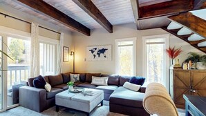 Una sala de estar con un sofá seccional, una mesa de centro y un rascador para gatos. Hay ventanas grandes y un mapa mundial en la pared.