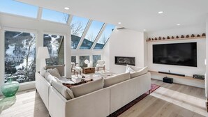 A modern living room with a large window, a sofa, a flat-screen TV, and a fireplace. - Luxury, designer mountain perch with amazing views, chef's kitchen & hot tub (Vail)