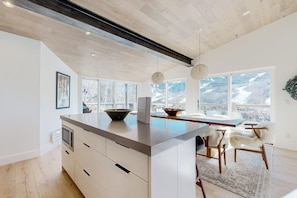 A modern kitchen with a large island, wooden flooring, and large windows offering a mountain view. - Luxury, designer mountain perch with amazing views, chef's kitchen & hot tub (Vail)