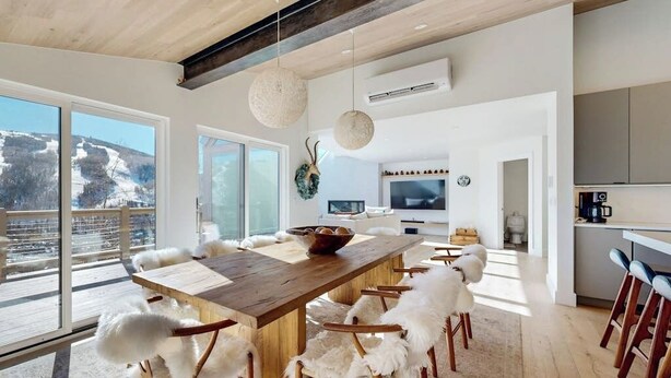 A modern interior with a wooden dining table, white furry chairs, a kitchen area, and large windows offering a view of a snowy landscape. - Luxury, designer mountain perch with amazing views, chef's kitchen & hot tub (Vail)