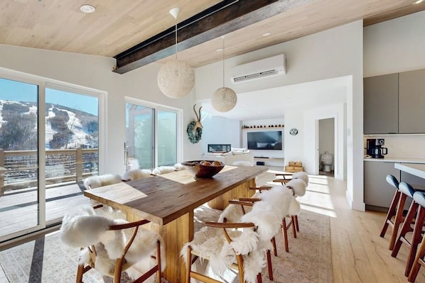 A modern interior with a wooden dining table, white furry chairs, a kitchen area, and large windows offering a view of a snowy landscape. - Luxury, designer mountain perch with amazing views, chef's kitchen & hot tub (Vail)