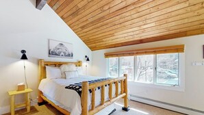 Une chambre à coucher avec un lit en bois, une table de chevet, une lampe, une fenêtre donnant sur un beau paysage et un plafond avec des poutres apparentes.