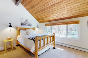 Ein Schlafzimmer mit einem hölzernen Bett, einem Nachttisch, einer Lampe, einem Fenster mit Blick und einer Decke mit Holzbalken.