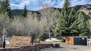 A sign for Timber Falls buildings with a stone wall, a wooden signboard, and a view of a lodge with a mountain backdrop. - Mountain view condo with 2 balconies, pool & pickleball - steps to free shuttle (Vail)
