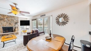 A living room with a stone fireplace, a ceiling fan, a dining table with chairs, and a large mirror on the wall. - Mountain view condo with 2 balconies, pool & pickleball - steps to free shuttle (Vail)