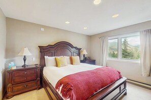 A bedroom with a large bed, two bedside tables, a window with curtains, and a view of greenery. - Charming townhouse with private hot tub, mountain views, and family-friendly (Vail)