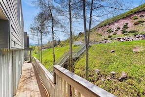 A wooden deck with a metal railing, a staircase leading to a grassy area, and a hillside in the background. - Conveniently located condo w/ full kitchen, W/D & balcony with great views (Vail)