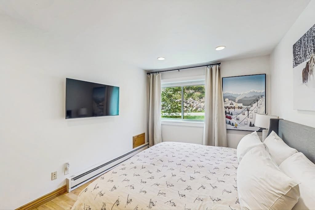 Un dormitorio con cama, televisor montado, ventana con cortinas y vista a la montaña.