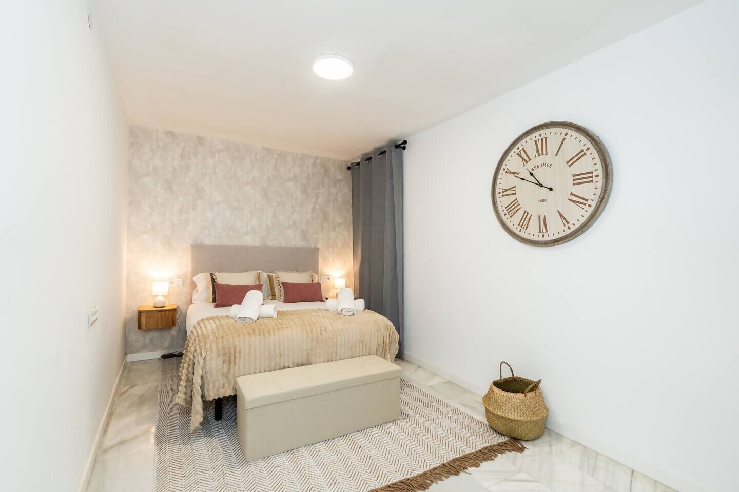 Un dormitorio con un reloj de pared grande, una cama con almohadas, una mesita de noche con lámpara y una cesta tejida en el suelo.