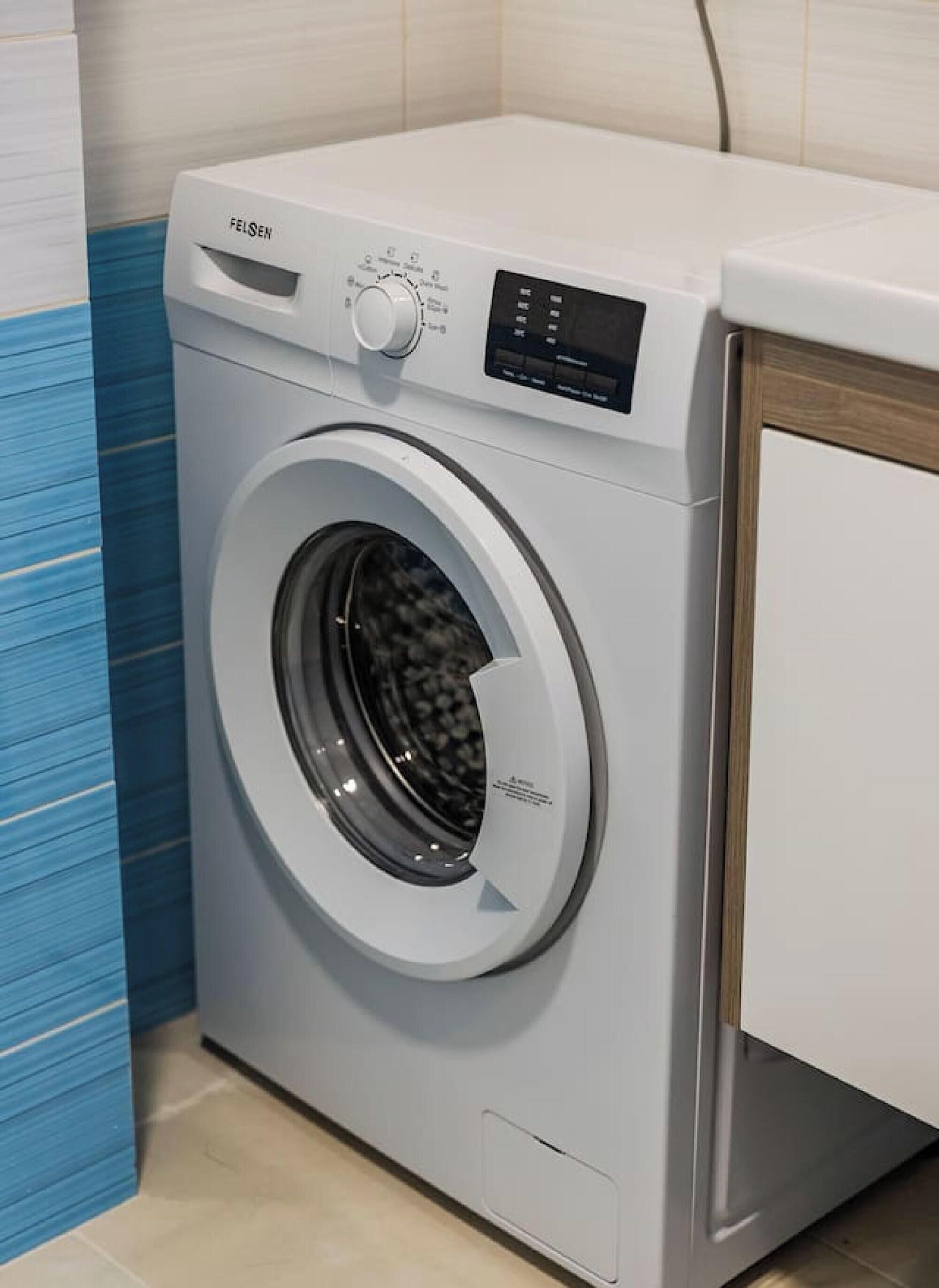 Lavadora de carga frontal con panel de control, ubicada en una habitación con piso de baldosas y pared de acento azul.