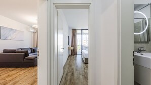 A modern hotel room with a wooden floor, a sofa, a bed, a bathroom with a mirror and sink, and a view of the city. - Adriatic Escape Apartment Steps From the Beach (Shengjin)