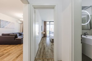 A modern hotel room with a wooden floor, a sofa, a bed, a bathroom with a mirror and sink, and a view of the city. - Adriatic Escape Apartment Steps From the Beach (Shengjin)