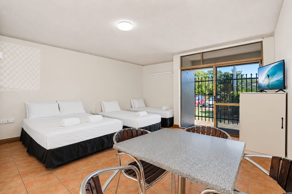 Habitación de hotel con dos camas, una mesa de comedor, sillas, televisor y puerta corrediza de vidrio que da a un balcón con vistas al exterior.