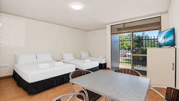 A hotel room with two beds, a dining table, chairs, a TV, and a sliding glass door leading to a balcony with a view of the outdoors.