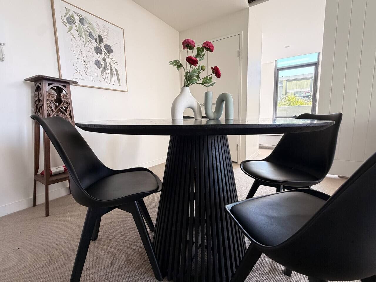 A modern dining area with a black table and chairs, a vase with flowers, and a framed botanical illustration on the wall.