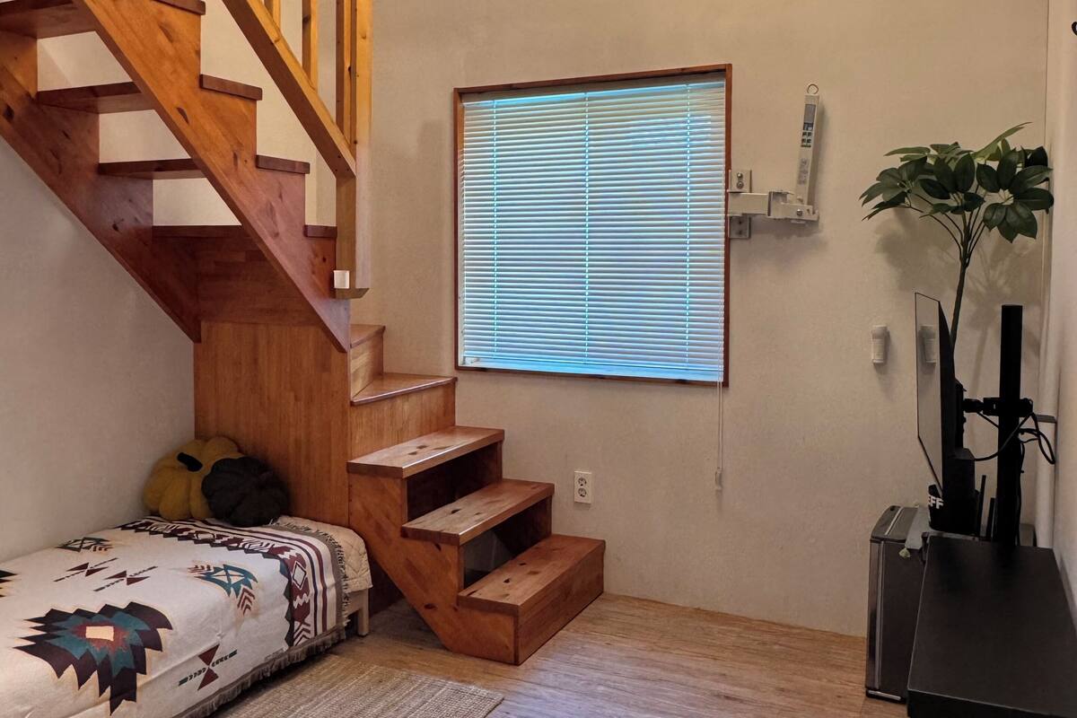Un escalier avec des rampes en bois, une fenêtre fixée au mur avec des stores, un luminaire suspendu et une petite plante.