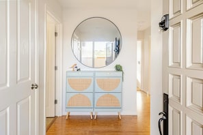 A hallway with a round mirror, a light blue cabinet with wicker doors, and a door with a security camera.