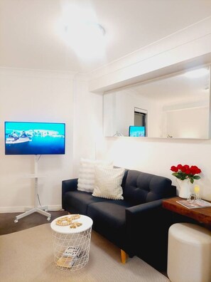 A modern living room with a dark sofa, a flat-screen TV, a round side table with a magazine, and a small white side table with a vase of red flowers. - Central Loft on Broadway + Parking/Pool (Glebe)