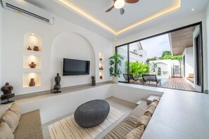 Ein modernes Wohnzimmer mit Flachbildfernseher, eingelassener Beleuchtung, Deckenventilator, großem Fenster und einer Sitzecke mit Sitzsack und Kissen.