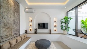 Una sala de estar moderna con un sofá, un pouf redondo, un televisor de pantalla plana, cerámica decorativa y una planta grande.