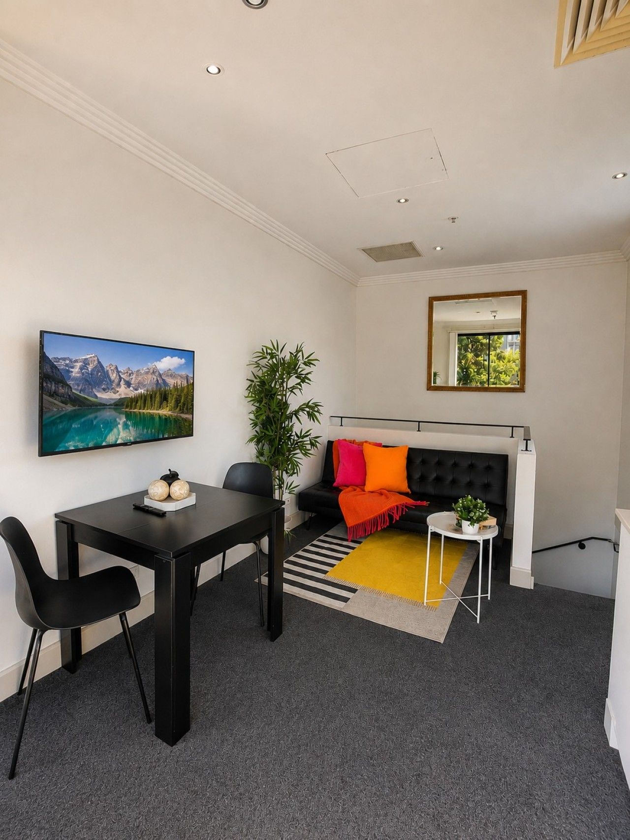 A modern living room with a black sofa, a dining table with chairs, a TV mounted on the wall, a potted plant, and a window with a view of greenery.