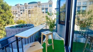 Un balcon avec une petite table et deux tabourets, une fenêtre et une vue sur un immeuble à l’architecture classique.