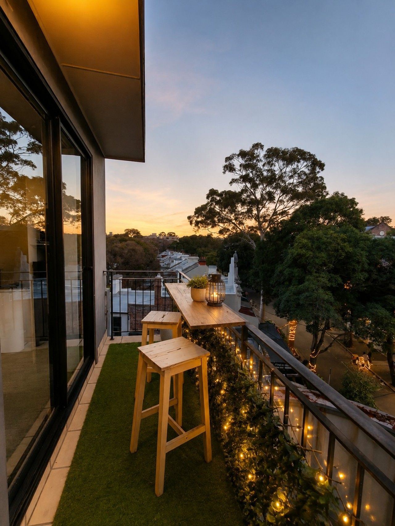 A balcony with a view of trees and buildings, a wooden table, and two wooden stools.