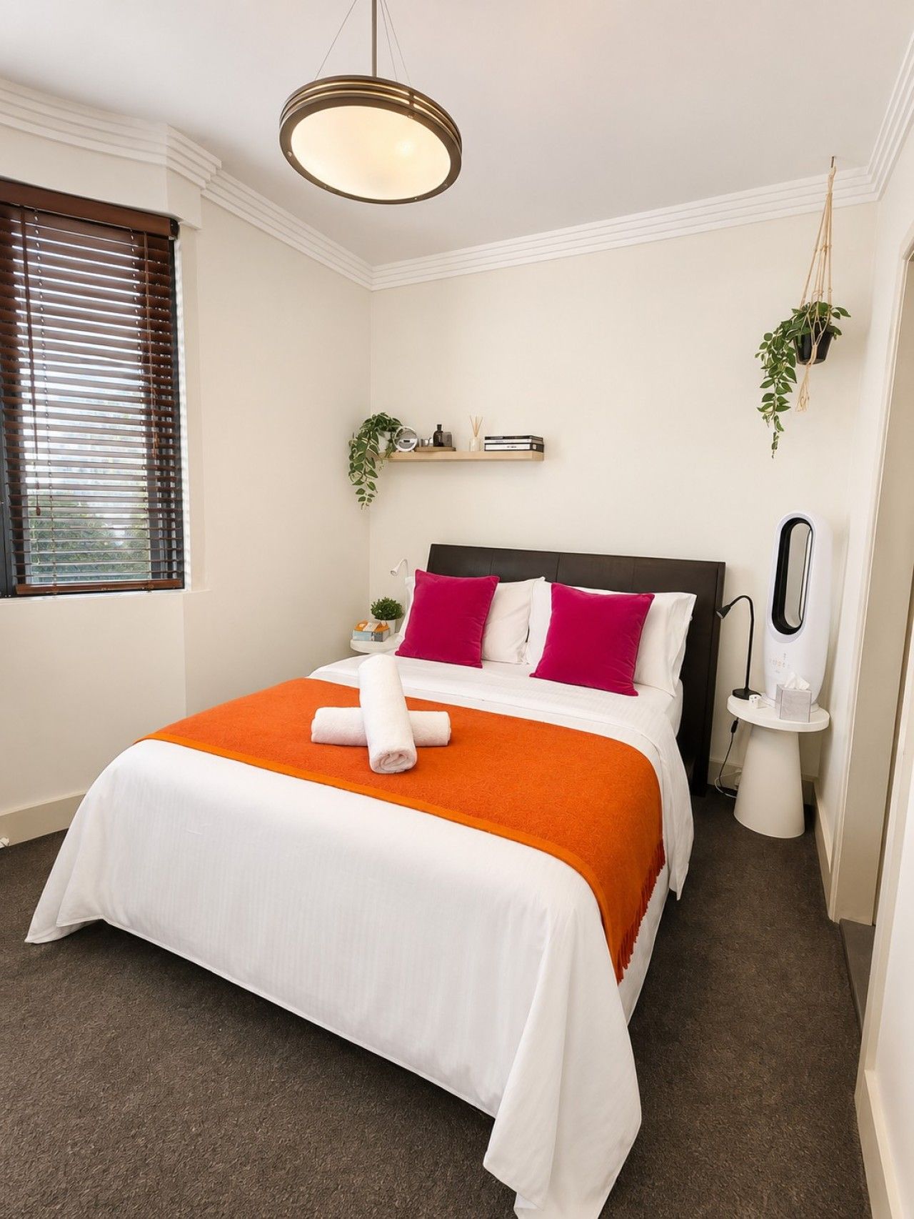 Una cama bien hecha con colcha blanca y naranja, cabecera y dos almohadas. Hay una mesita de noche con una lámpara y una planta pequeña.