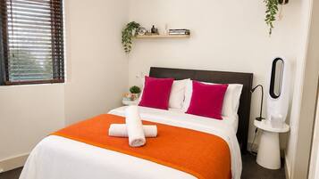 Una cama bien hecha con colcha blanca y naranja, cabecera y dos almohadas. Hay una mesita de noche con una lámpara y una planta pequeña.