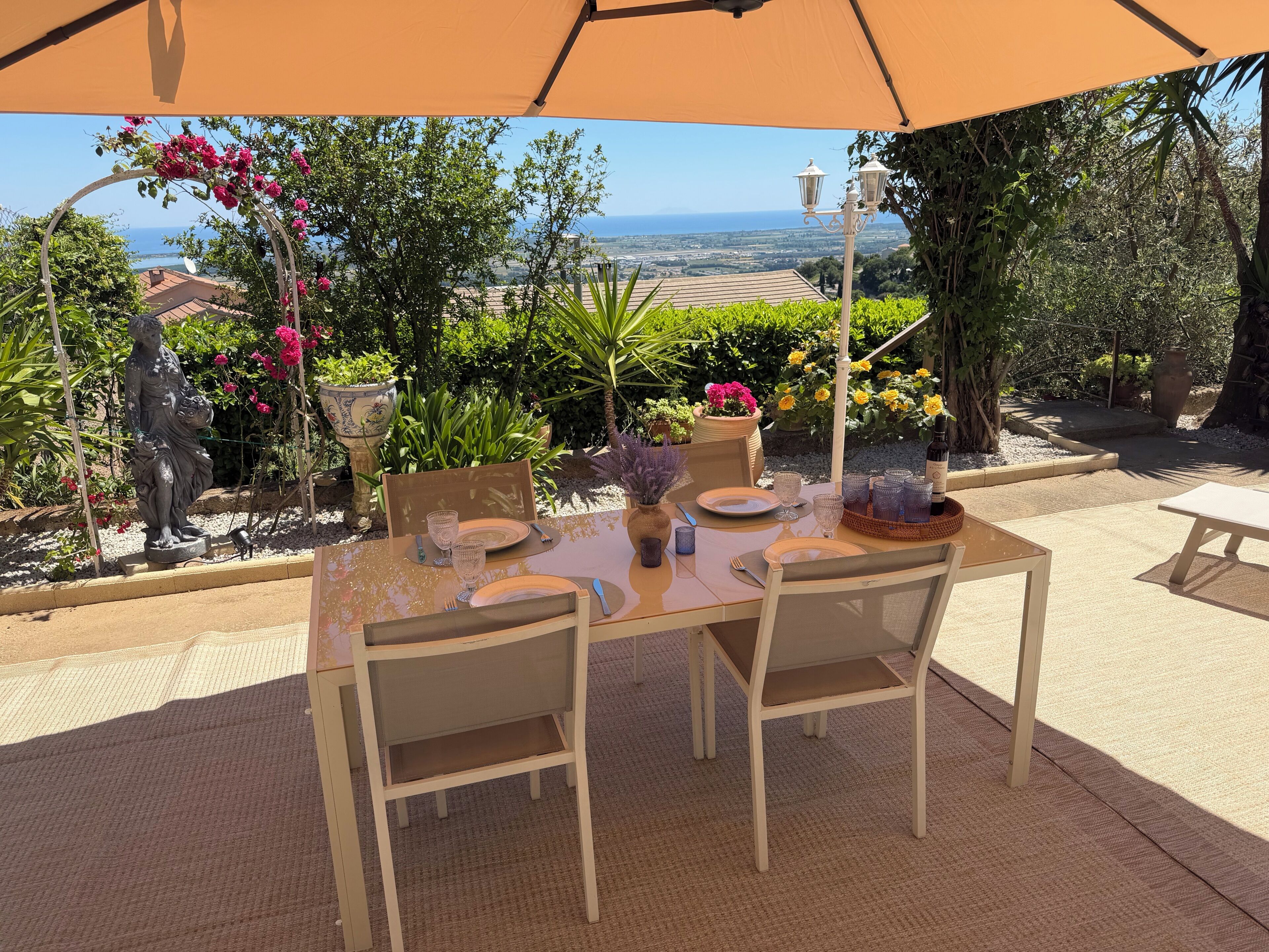 An outdoor dining area with a table set for four, surrounded by greenery and a view of the sea.