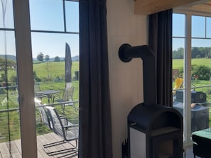 Ein Zimmer mit großen Fenstern, einem schwarzen Kaminofen und Blick auf ein grünes Feld.