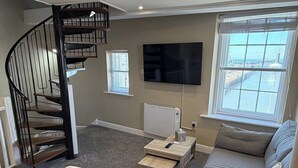 Una sala de estar con un sofá gris, una mesita, un televisor montado en la pared, una ventana con persianas y una escalera de caracol.