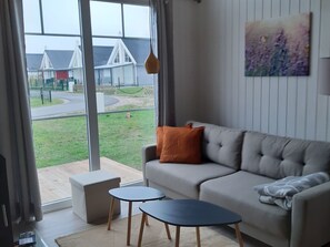 Ein Wohnzimmer mit einer Couch, zwei kleinen Tischen, Blick auf eine Wohngegend durch große Fenster und einem Bild an der Wand.