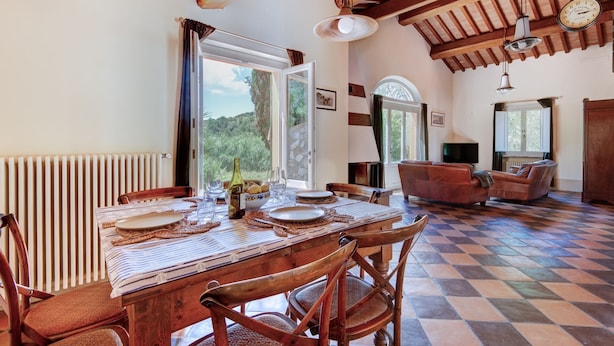 Uma sala de jantar com uma mesa posta para quatro pessoas, um sofá e uma televisão, num ambiente com vigas de madeira e chão xadrez.