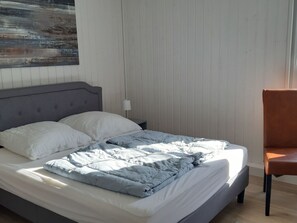 Ein Bett mit grauem Kopfteil, weißen Kissen und einer grauen Decke. Ein Nachttisch mit Lampe und ein brauner Stuhl stehen daneben.