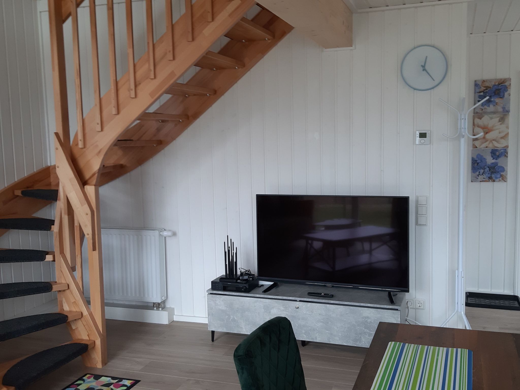 A living room with a wooden staircase, a flat-screen TV, a dining table, and a clock on the wall.