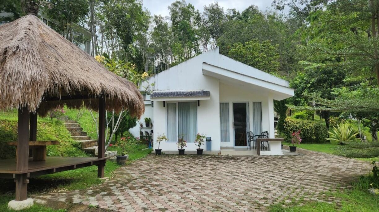 A white building with a thatched structure nearby, surrounded by lush greenery.