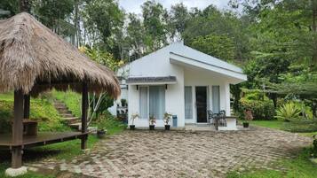 A white building with a thatched structure nearby, surrounded by lush greenery.