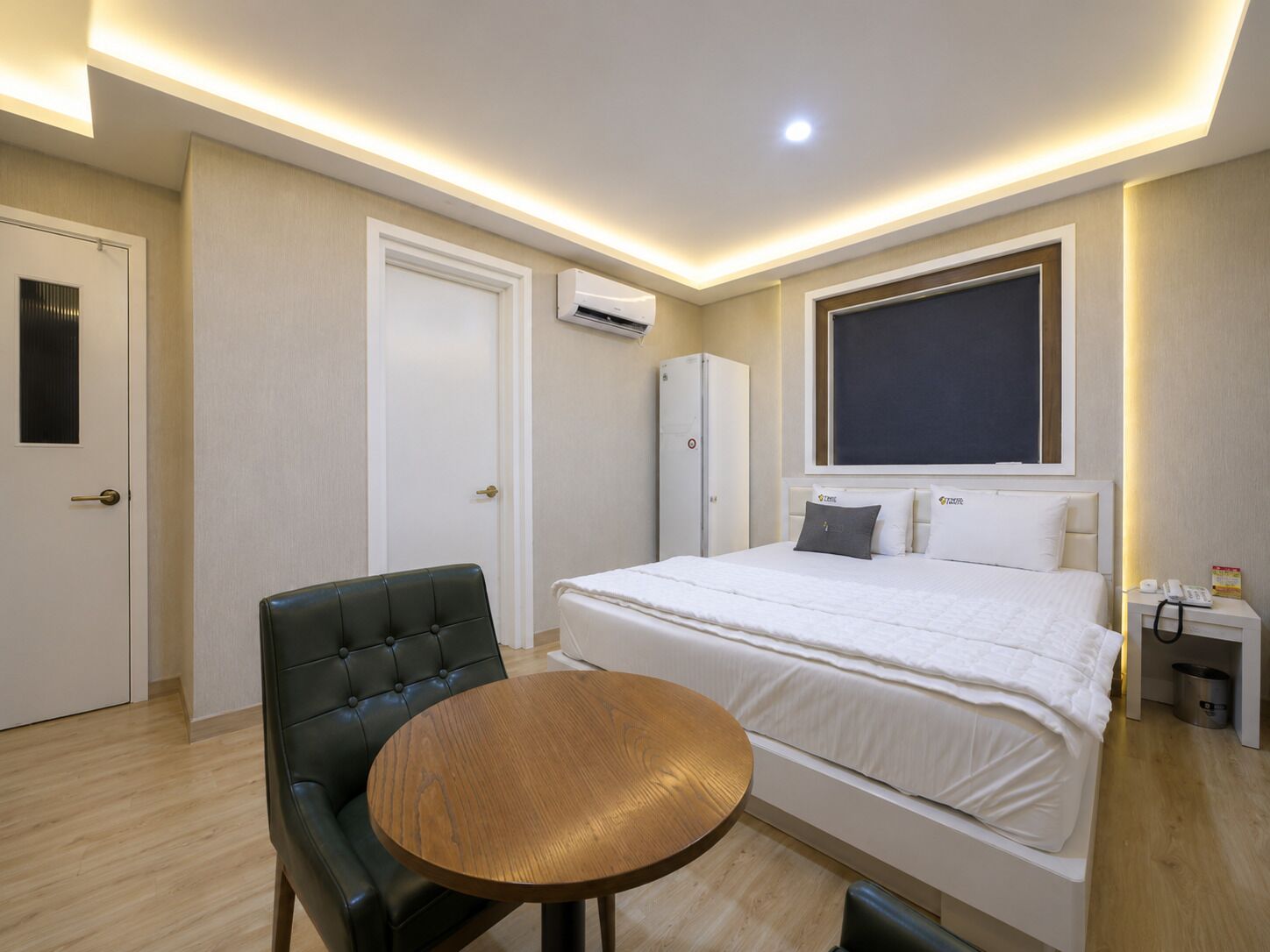 A hotel room with a bed, a round wooden table, a black leather chair, a wardrobe, an air conditioning unit, and a bedside table with a phone and a trash bin.
