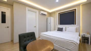 A hotel room with a bed, a round wooden table, a black leather chair, a wardrobe, an air conditioning unit, and a bedside table with a phone and a trash bin.