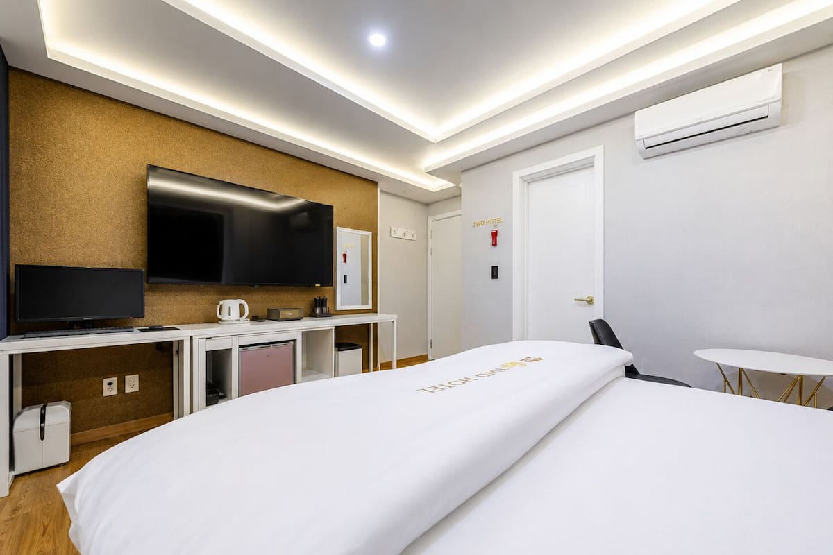 A hotel room with a bed, a desk with a TV, a coffee maker, a mini fridge, and an air conditioning unit.