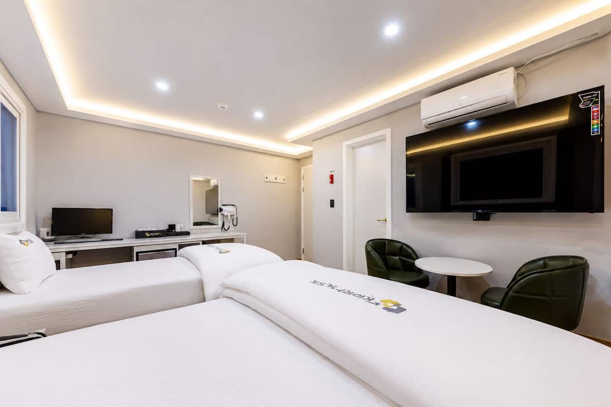 A hotel room with two beds, a desk with a flat-screen TV, a small round table, and a green chair.