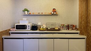 Una kitchenette con microondas, tostadora y un refrigerador pequeño. Hay repisas arriba con varios objetos y una cámara en la pared.