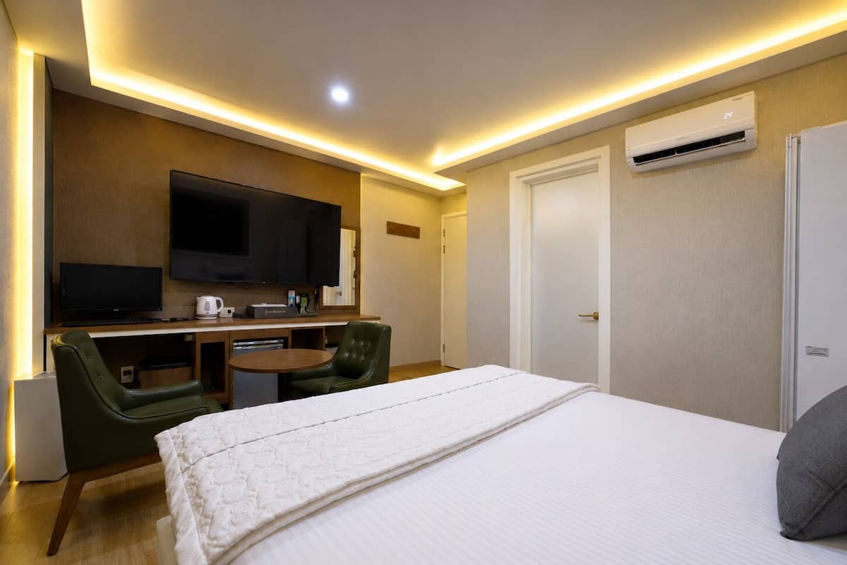 A hotel room with a bed, a desk with a flat-screen TV, a green chair, and an air conditioning unit.