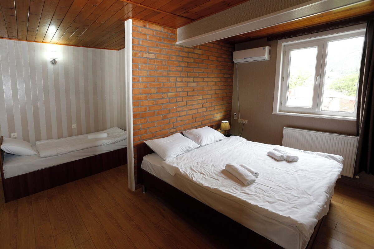 A bedroom with a wooden floor, a brick wall, a bed with white linens, a window with curtains, and a radiator.