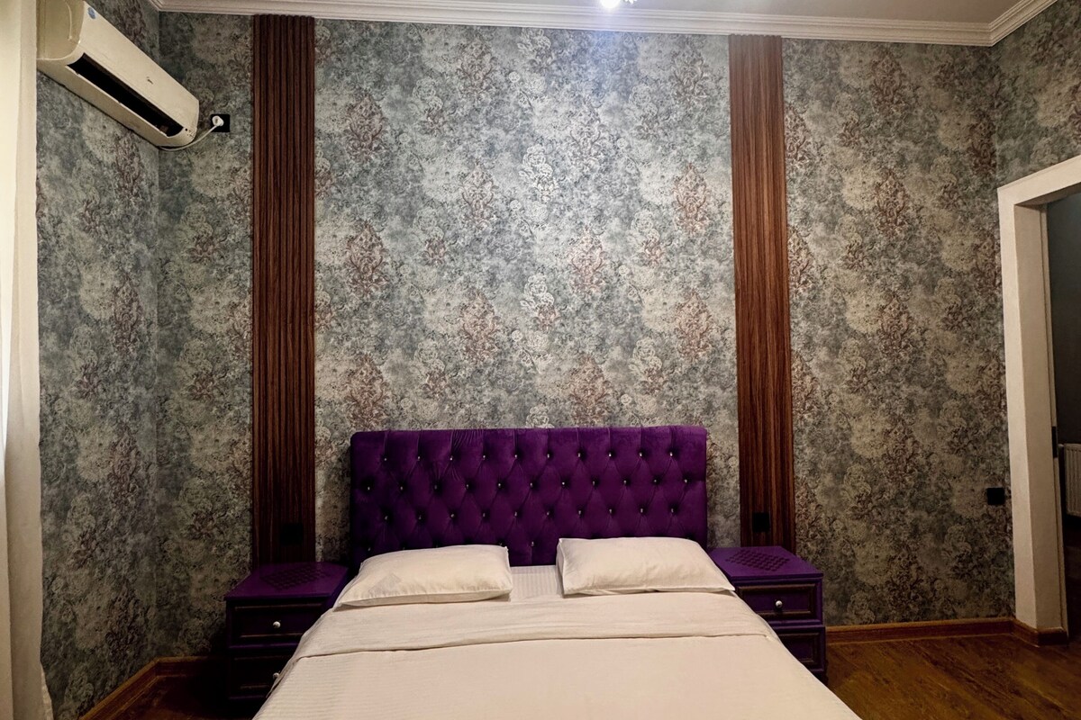 A bedroom with a large bed, a chandelier, a wall air conditioner, and patterned wallpaper.